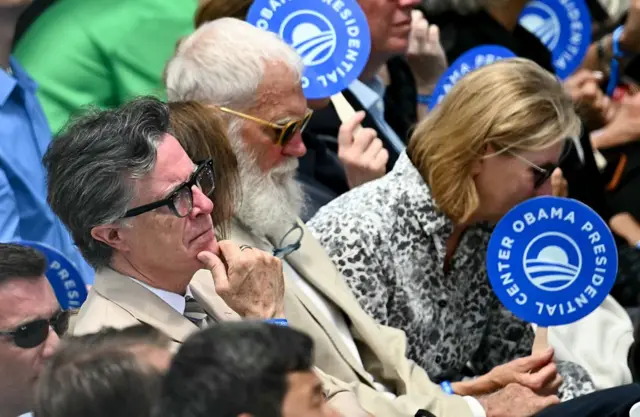 Stephen Colbert y David Letterman siguen el evento rodeados de más personas. Algunas de ellas portan unos carteles circulares del Obama Presidential Center