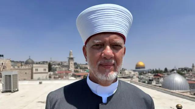 Un hombre con barba y túnica gris de pie en una azotea en Jerusalén, con la cúpula dorada de la mezquita de Al Aqsa al fondo.