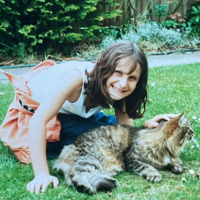 Una niña en el jardín con un gato atigrado de pelo largo. Está sonriendo a la cámara y tiene el pelo largo y castaño. Lleva vaqueros azules y una blusa blanca sin mangas con un chándal naranja atado alrededor de su cintura.