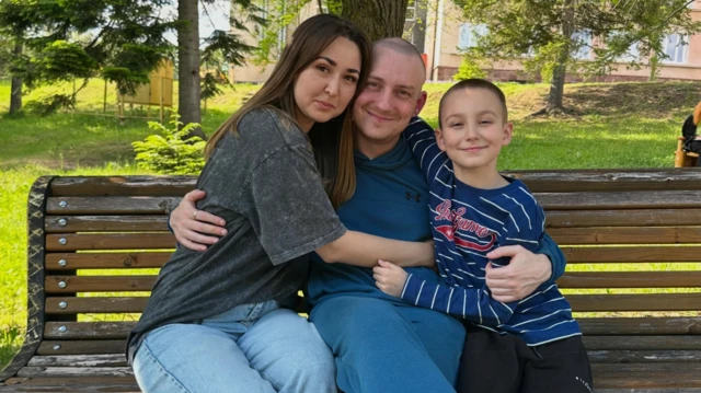 Con un jersey y pantalones azules, el mayor Oleksandr Ivanov está sentado en un banco del parque entre su esposa, Nelly, con una camiseta gris antracita y unos vaqueros azules, y su hijo pequeño, Demid, con pantalones negros y una camiseta azul a rayas con letras rojas, todos sonriendo y abrazándose.