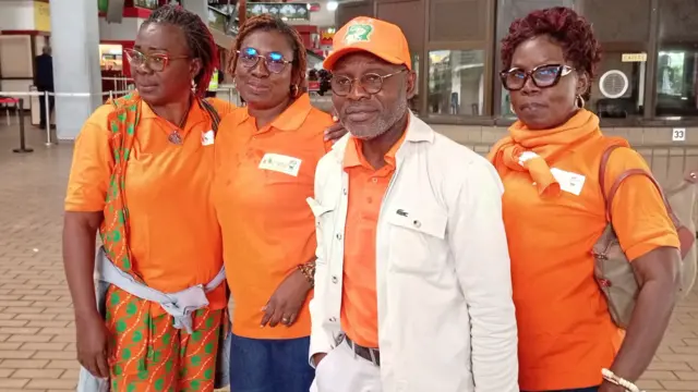 Un hombre que lleva una camiseta naranja, una gorra de béisbol y una chaqueta blanca mira a la cámara. Lleva gafas y tiene barba corta. Junto a él hay tres mujeres, que también llevan camisetas polo naranjas.