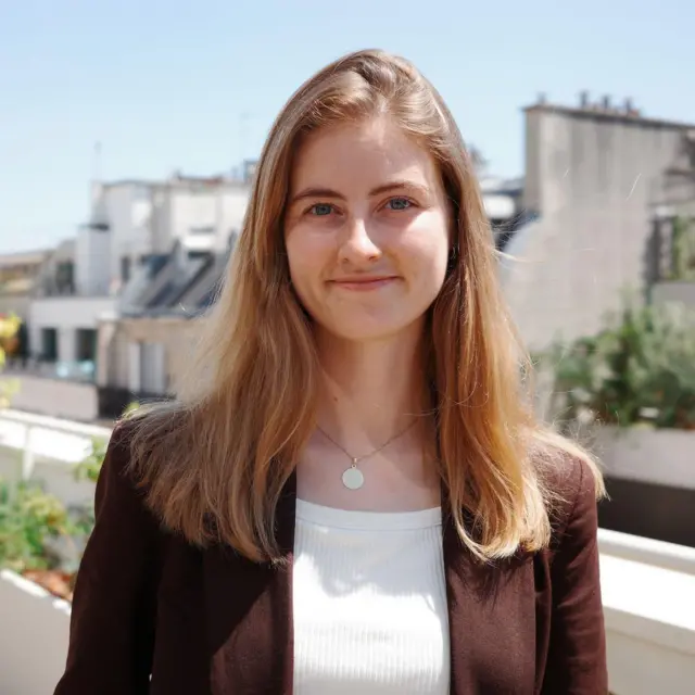 Emma Fourreau está de pie en un balcón soleado, con plantas de fondo. Lleva una chaqueta marrón abierta y, debajo, un top blanco de canalé. Emma Fourreau sonríe en la imagen.