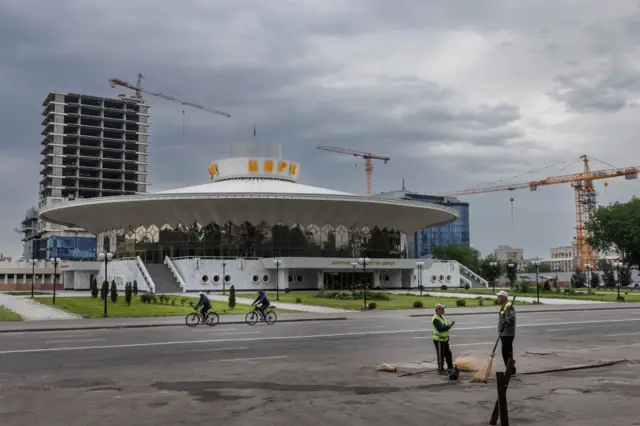 Un edificio con forma de "plato volador" en el centro de Biskek. En el tejado se lee "Circo" en letras grandes. Al fondo se ven tres grúas de obra y un rascacielos en construcción.