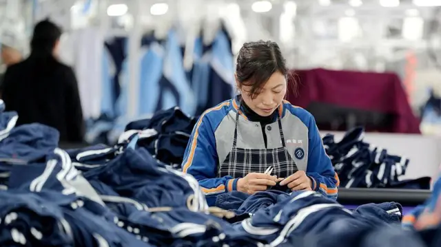 Una mujer cose un trozo de tela entre muchas prendas en lo que parece ser una fábrica. Lleva un uniforme y un delantal estampado