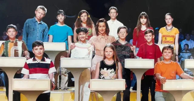 Con una camiseta roja, Oleksandr Ivanov es uno de los 13 niños y niñas que están de pie, en tres filas, detrás de atriles blancos, frente al público de un estudio de televisión sentado en gradas. Una presentadora adulta, vestida con una blusa color melocotón, está de pie en el centro, abrazando a dos de los niños.