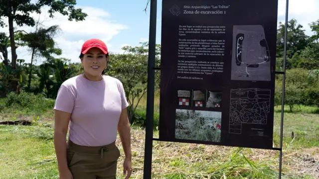 Mujer junto a un cartel informativo de un sitio arqueológico en un entorno rural.