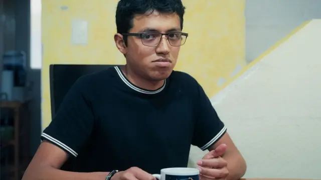 Julian lleva puesta una camiseta azul marino y blanca, y sostiene una taza de té; lleva anteojos y tiene el pelo oscuro.
