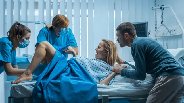 Una mujer dando a luz en un hospital acompañada de su pareja que le toma la mano, mientras dos parteras la asisten