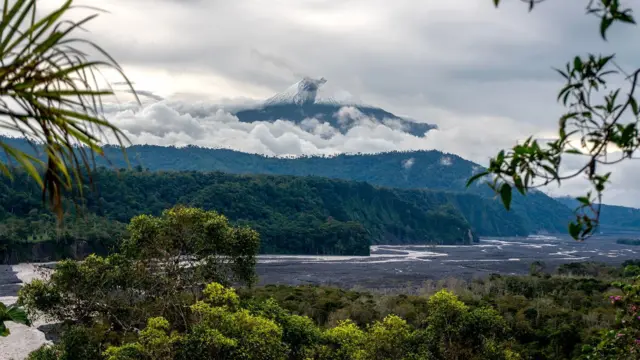 Volcán cubierto de nubes sobre un valle selvático con río serpenteante.
