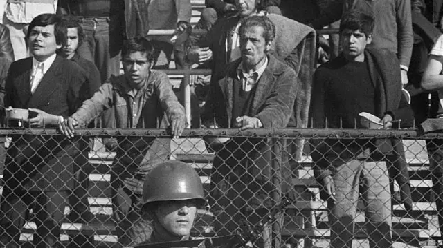 Fotografía en blanco y negro en la que se ve a personas detenidas en un estadio deportivo tras el golpe de Estado. Un soldado armado con un rifle se encuentra de pie frente a la valla, mientras los prisioneros esperan a ser interrogados.