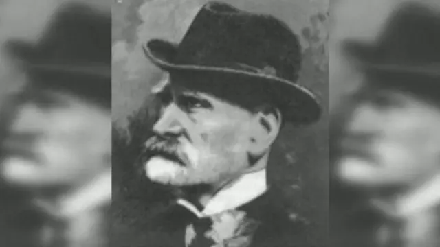 Foto en blanco y negro de Cobb Morley, de perfil, con un sombrero, camisa de cuello alto, corbata antigua y chaqueta, estilo victoriano. lleva barba y bigote. 
