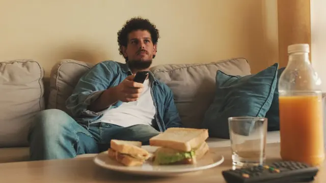 Un hombre con un control remoto sentado frente a una mesa con un sándwich en un plato, un vaso y una botella con jugo