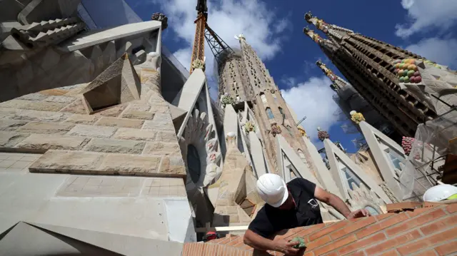 Un obrero con casco blanco y camiseta negra en trabajos de construcción con varias torres elevándose a sus espaldas.