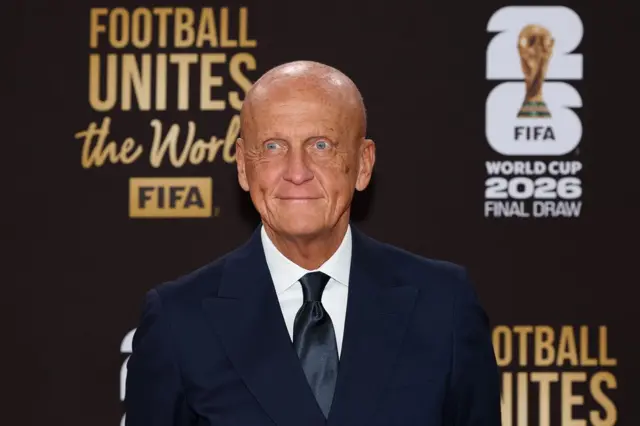 Pierluigi Collina, vestido de traje negro y corbata negra con una camisa blanca, está sentado en el Centro John F. Kennedy. Atrás tiene una pancarta que dice "Football Unites The World. FIFA World Cup 2026 Final Draw" y tiene una foto de la Copa del mundo