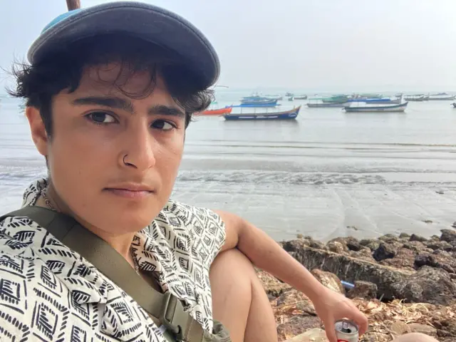 Sophia se sienta en la playa en una foto de selfie. Muestra una hilera de barcos que se balancean en el horizonte, mientras ella está sentada con una camisa estampada, una gorra de béisbol y sostiene una lata en la mano.