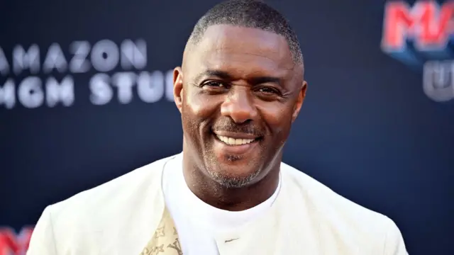 Luther Idris lleva una chaqueta color crema y una camiseta blanca