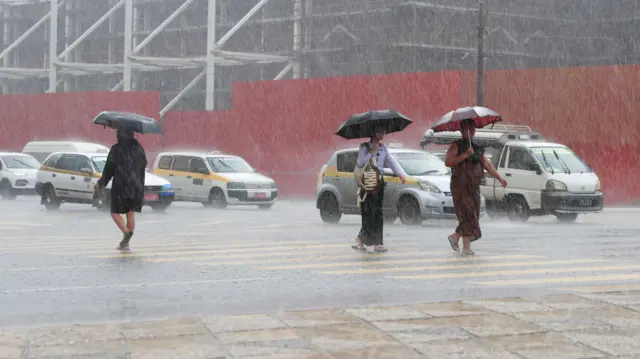 Tres personas cruzan una carretera bajo una lluvia intensa en Myanmar, con tráfico al fondo. Todas llevan paraguas mientras la lluvia rebota en el paso de peatones.