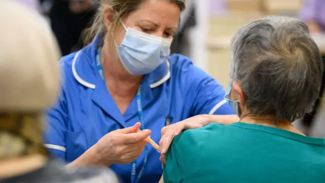 La imagen muestra un entorno sanitario en el que un profesional de la salud administra una inyección en la parte superior del brazo de otra persona. El profesional viste un uniforme azul y una mascarilla, y sostiene una jeringa con cuidado mientras prepara o aplica la inyección. La persona sentada lleva la manga ligeramente subida para dejar al descubierto la zona donde se administrará la inyección. El fondo aparece suavemente desenfocado, con otras personas y elementos que sugieren una clínica o un centro de vacunación con mucha actividad.