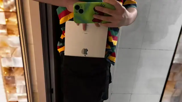 Una mujer vestida con ropa masculina de talla grande sostiene un teléfono móvil en la mano izquierda, toma una foto mientras guarda una tableta portátil en el bolsillo con la otra mano. Su camiseta es multicolor y lleva pantalones negros