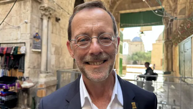 Un hombre con gafas pequeñas de montura metálica redonda, traje gris, camisa blanca y un pequeño distintivo dorado en la solapa sonríe en primer plano. En el fondo se observa una barrera.
