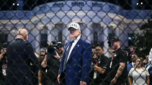 El presidente de los Estados Unidos, Donald Trump, visto a través de una cerca de alambre metálico. Lleva un traje azul y una gorra de béisbol blanca que dice "EE. UU.". Al fondo hay una multitud de personas y la Casa Blanca