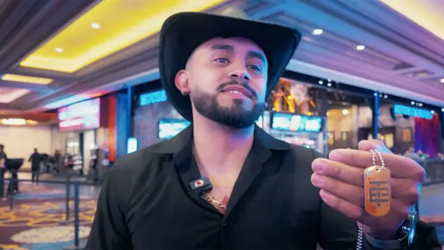 Un fan de El Temach lleva una camisa negra de cuello abierto y un sombrero Stetson negro, y sostiene una cadena especial que le compró al influencer como mercancía; es de color naranja con un patrón geométrico gris.