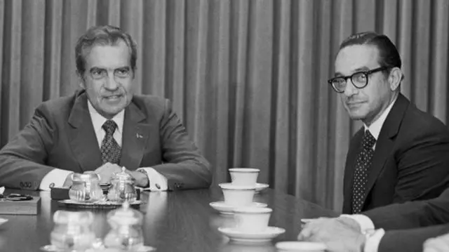Una fotografía en blanco y negro de Richard Nixon y Alan Greenspan. Están sentados ante una mesa cubierta de tazas de té.