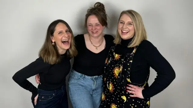 Tres mujeres de unos 30 años riendo y abrazándose.