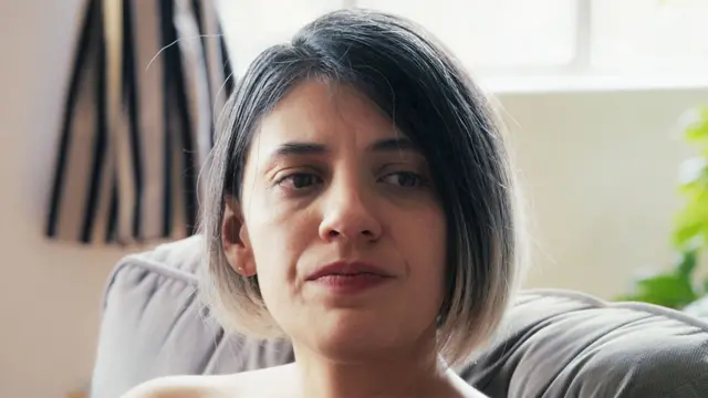Alex lleva el cabello corto, tiene los ojos oscuros, lleva pintalabios rojo, está sentada en un sofá y tiene un aspecto apesadumbrado.