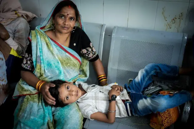 Yash, de seis años, recostado en el regazo de su madre, en sillas que parecen ser de la sala de espera del hospital. La madre viste un sari verde y azul, él, jeans azules y camisa de manga corta gris y blanca con diseños geométricos.