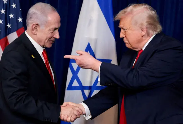 Benjamin Netanyahu y Donald Trump se estrechan la mano mientras están de pie frente a las banderas de Estados Unidos e Israel. Ambos visten trajes azul oscuro con corbatas rojas, mientras Trump señala a Netanyahu.