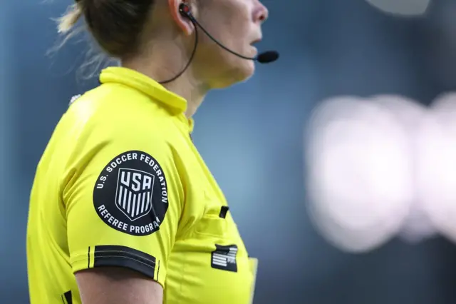 El perfil de una árbitra que lleva el cabello recogido y tiene un audífono con micrófono. Lleva un uniforme amarillo y en el brazo tiene el escudo que dice U.S. Soccer Federation, USA, Referee Program