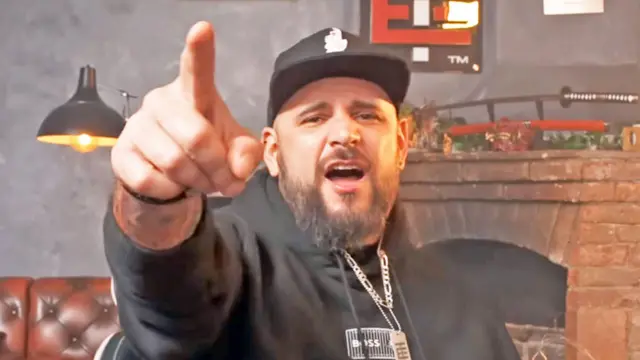 El Temach lleva una sudadera negra con capucha y una gorra blanca y negra, y tiene barba; está señalando y gritando a la cámara.