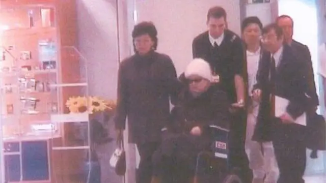 Ko Yong-hui, con gafas de sol y un gorro blanco, está sentada en una silla de ruedas que empuja un hombre. Hay varias personas a su alrededor en un hospital de Francia.