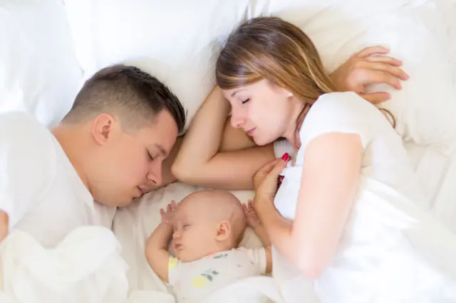 Un padre y una madre dormidos con su bebé en la misma cama. Están cubiertos de sábanas blancas y recostados en almohadas blancas