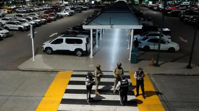 Miembros de las fuerzas armadas de Ecuador montan guardia tras un ataque armado en el aeropuerto de Guayaquil. Se observan dos soldados armados y otros tres hombres uniformados de pie en un paso de peatones que conduce al estacionamiento.