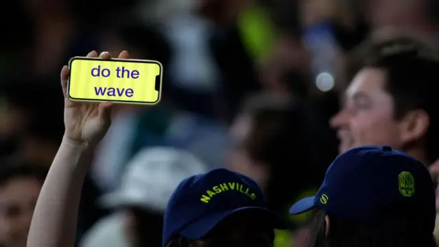 Brazo levantado con la mano sosteniendo un iPhone en cuya pantalla se ve escrito "do the wave" en letras azules y fondo amarillo. De la dueña sólo se ve su cachucha con la palabra Nashville, y la de otra persona a su lado, y en el fondo se ven otras personas borrosas. 
