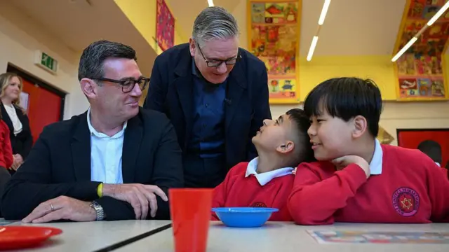 Keir Starmer mira por encima del hombro de Andy Burnham mientras charlan con dos niños con uniforme escolar durante una visita a un club de desayuno en Gran Manchester, en abril.