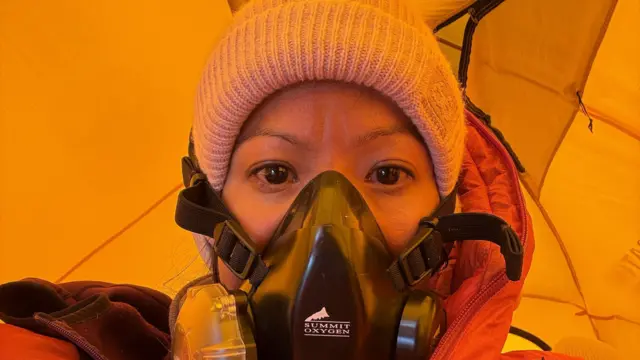 Purnima está de pie dentro de una tienda de campaña de color amarillo brillante, vistiendo un traje de protección climática rojo para gran altitud y una máscara de oxígeno negra.