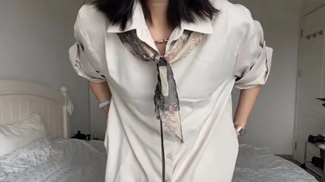Una mujer con el pelo hasta los hombros, vestida con una camisa grande de color blancuzco, una bufanda negra y crema con estampados alrededor del cuello, parada frente a una cama