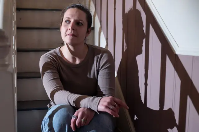 Ruth está sentada en una escalera interior, iluminada por una luz lateral que proyecta una sombra nítida sobre una pared revestida de paneles; mira ligeramente hacia un lado con un brazo apoyado en la rodilla levantada y viste una prenda de manga larga y vaqueros.