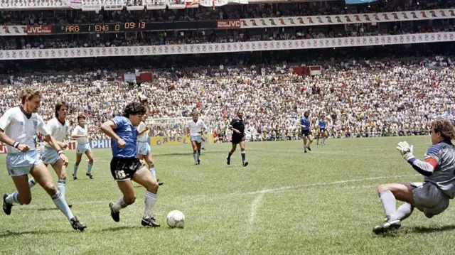 Se ve a Maradona regateando el balón a través de varios futbolistas ingleses y se ve al portero inglés lanzándose hacia un lateral.
