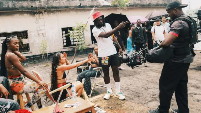 Meji Alabi, con un sombrero rojo, una camiseta blanca, pantalones cortos negros y calcetines y zapatillas blancas, sostiene una cámara y señala durante una sesión fotográfica. A su lado hay mujeres sentadas en tumbonas junto a un árbol delgado. Un camarógrafo, con una gorra negra y vestido con ropa oscura, está de pie al otro lado y el personal de producción, algunos con sombrillas negras, está detrás.
