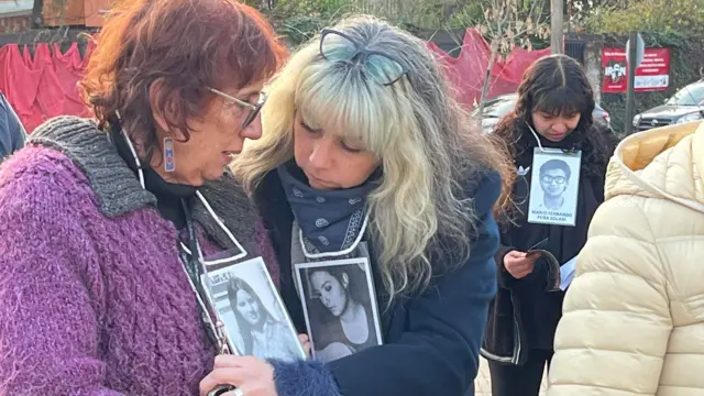 En la imagen, se ve a Holzapfel, a la izquierda, junto a otra mujer rubia a su derecha. Las mujeres se encuentran frente al monumento conmemorativo y llevan colgadas al cuello fotografías en blanco y negro de personas desaparecidas y antiguas víctimas del régimen de Pinochet.