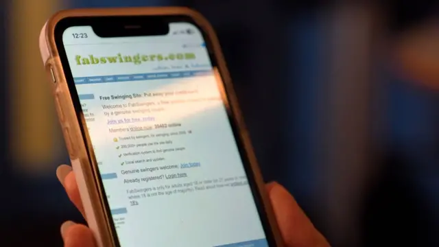Primer plano de un teléfono inteligente que muestra la página de inicio del sitio web FabSwingers, sostenido en una mano, con la pantalla ligeramente desenfocada sobre un fondo con poca iluminación.