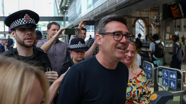 Andy Burnham, flanqueado por dos agentes de policía. Detrás de él, una multitud de personas le toma fotos.