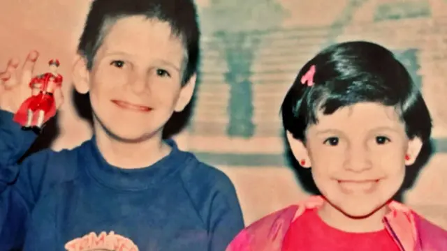 Un niño con una camiseta azul y una niña con una camiseta rosa, ambos de cabello oscuro, sonríen a la cámara; el niño sostiene una figura de juguete.