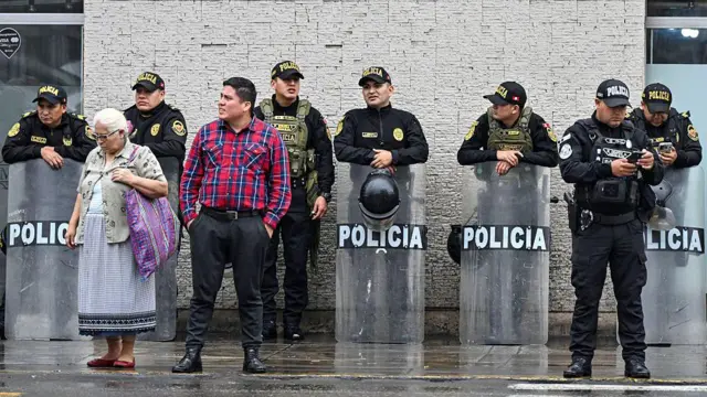 Una señora con un vestido azul y un bolso morado al lado de un hombre con una camisa a cuadros y un pantalón gris están parados en la calle. Atrás de ellos hay una fila de funcionarios de la policía, algunos con escudos, que hacen guardia al Palacio de Gobierno en Lima el 3 de junio de 2026