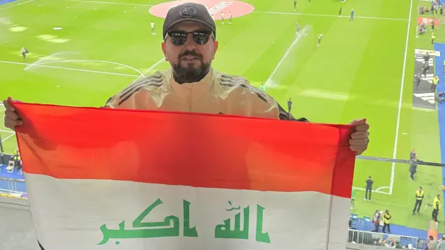 Un hombre con una gorra de béisbol oscura y gafas de sol sostiene una bandera iraquí en un estadio. Detrás de él se ve un campo de fútbol.