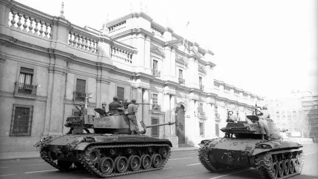 Foto de archivo de 1973: La fotografía en blanco y negro muestra dos tanques militares acercándose a un gran edificio administrativo, con soldados apuntando con sus rifles y listos para disparar.